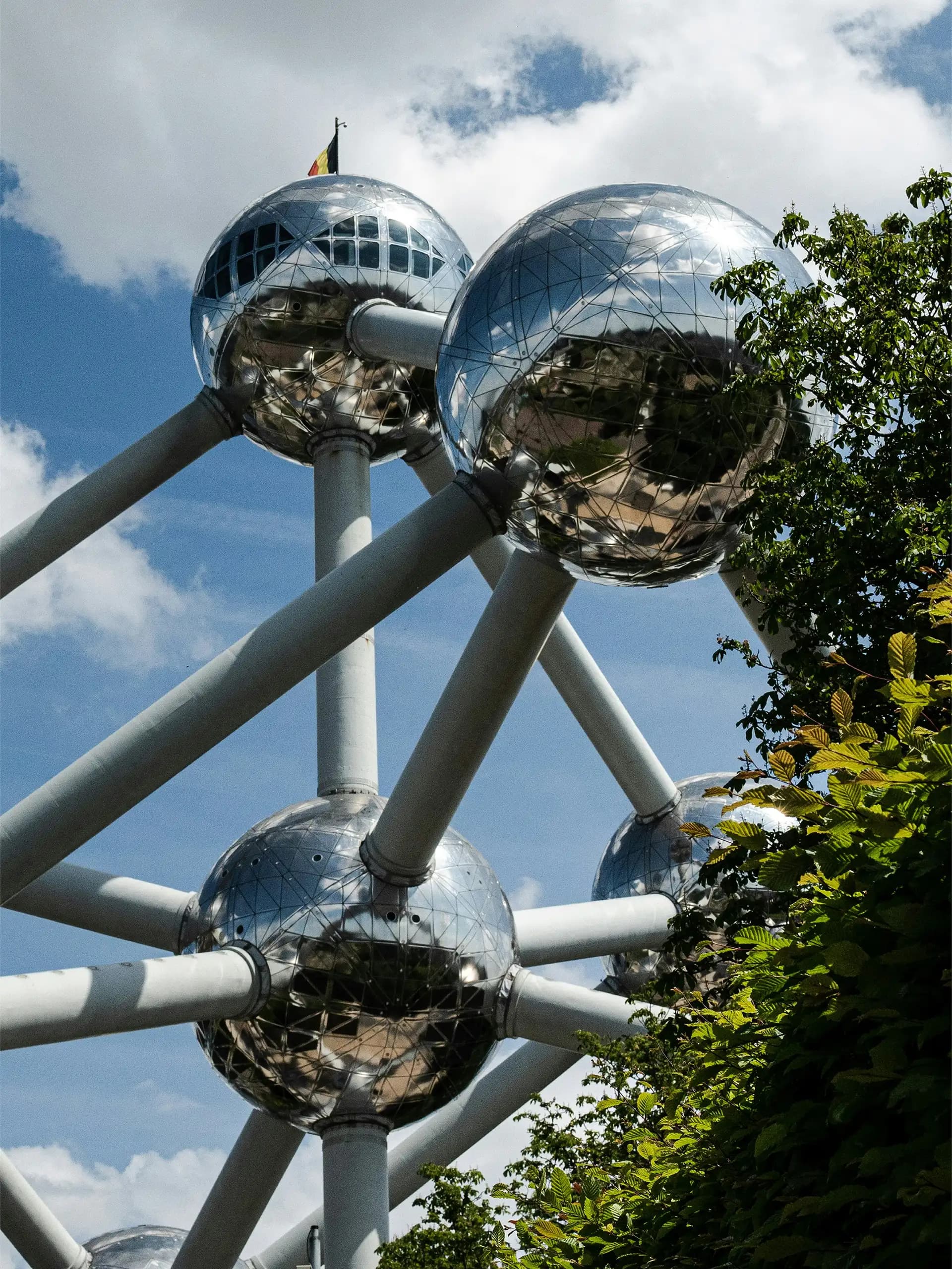 Atomium i Bryssel – blanka, sfäriska stålkulor sammanlänkade av rör, fotograferade underifrån mot en blå himmel med moln och gröna träd i förgrunden.
